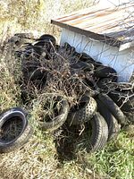 Pile of used tires including car and ATV tires stacked outdoors beside a metal roofed shed with weeds growing around them.