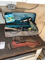 Metal toolbox open showing contents: electric drill, glue stick applicators, pipe wrenches, wire brush, and toolbox itself.
