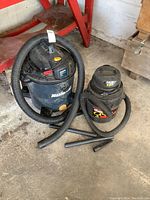 Two shop vacuums side by side: Mastercraft 10 gallon and smaller 4 gallon Shop Vac, both with hoses and vacuum attachments on floor beside them.