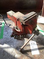 Side view of red 5 inch bench vise showing handle and jaws, placed on concrete surface with shadows.