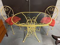 View of entire table and two chairs showing yellow wrought iron frames and oval glass tabletop with red cushions on chairs.