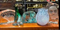 Four glass items on wooden surface including an etched iceberg glass paperweight, a crystal owl paperweight, a mottled purple-white bud vase, and a green-blue glass duck figurine, all visible and arranged side by side.
