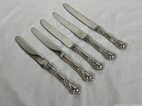 Full view of five dinner knives laid flat showing entire knife length, handles have ornate sterling silver detail and blades are steel