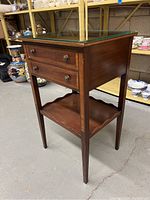 Side view of antique 2-drawer side/accent table with glass top showing wood grain and inlaid maple and birch borders, round metal drawer knobs, and lower scalloped shelf.