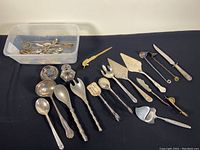 Full assortment of vintage serving utensils arranged on black fabric backing with plastic bin containing some small items.