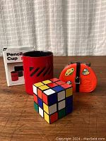 Overview photo showing desk items: red pencil cup, orange novelty bank resembling a suitcase, and partially scrambled Rubik's Cube puzzle toy