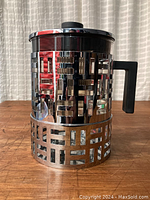 Closed view of the Bodum Eileen glass teapot with its pierced stainless steel sleeve and black plastic handle on a wood surface with curtain background.