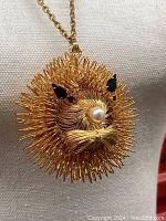 Close-up of the goldtone lion head pendant showcasing bent wire filigree, black rhinestone eyes, and pearl nose detail.