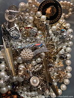 Top-down view of pile of assorted faux pearl necklaces and numerous brooches in mixed metals with rhinestones and other embellishments. Variety of shapes and sizes visible.