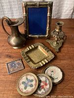 Brass decor lot showing picture frame, pitcher, nightstick, ashtray, and four coasters with needlework linings on wooden table.