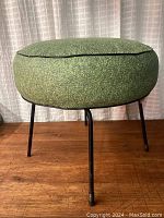 Side view of round green vinyl cushioned footstool showing black piping and three metal legs on wood floor against curtained background.