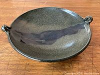 Top-down and angled views of a round pottery bowl with three sculptural handles and a gray glaze with a darker brushstroke streak through the center, placed on a wooden table.
