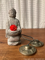 Front view of cement Buddha statue seated with bowl holding red flower, alongside Tingsha cymbals on wooden surface.