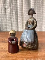 Front view of the two pottery figurines on wooden table with curtain background. The larger female figurine wears a blue pointed hat and apron, and the smaller child figurine in maroon dress holds a small white object.