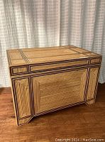 Rectangular rattan chest with dark cane geometric patterns on front and sides, showing minor wear on surface and wood grain on table below.