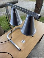 Side angle photo showing the aluminum heat lamps with perforated holes, attached to a rectangular metal stand sitting on a wooden table outdoors.