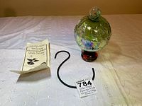 Photo showing the globe-shaped hummingbird feeder with iridescent glass, red flower feeding ports, the black metal S-hook, and the instruction leaflet on a white fabric surface