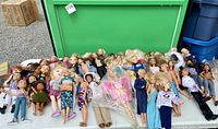 Collection of assorted Barbie, Ken, and other fashion dolls arranged on a table, showing variety in hair color and clothing styles.