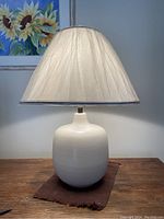 White ceramic lamp with rounded base and off-white fabric shade shown lit from above on wooden surface with framed floral painting background.