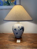 Full view of ceramic lamp on wooden surface showing beige fabric lampshade and ceramic base with blue grape and leaf design.