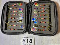 Top-down view of 12 multicolored spinner bait spoon fishing lures with gold hooks stored in a black foam insert inside a zipped case.