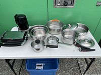Photo showing entire set of items on table, including panini toaster, kettle, various pots, lids, and metal bowls.