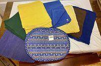 Photo showing assorted fabric placemats in various colors including yellow, blue, green, purple, brown and a blue floral patterned oval placemat, displayed on a table.