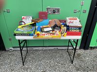 White foldable table with various board games including Scruples, Scattergories, Backgammon, Checkers, Careers, plus wooden paddle ball paddles, plastic paddle ball game, and golf balls.