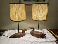 Pair of carved wooden duck lamps with shades, shown side by side on a white cloth.