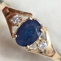 Close-up of 10K gold ring with oval sapphire center and two small diamonds on either side, showing facet detail and gold prong settings.