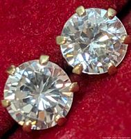 Close-up front view of two round clear cubic zirconia stones in 10K gold 6-prong settings, seated on red fabric background to show sparkle and detail.