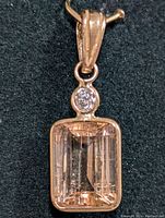 Close-up of the light peach rectangular faceted tourmaline set in 14K gold with a single round diamond above it on a black background.