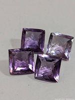 Four square faceted genuine amethyst gemstones with purple coloration placed on a light surface.