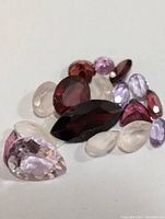 Close-up photo of assorted faceted genuine gemstones in various colors including deep red, light pink, purple, and clear, showing different cuts such as pear, oval, round, and marquise.