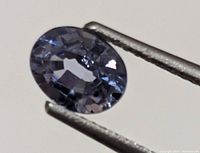 Close-up of oval cut Ceylon sapphire held by tweezers showing blue-violet facets and transparency.