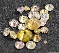 Multiple loose diamonds scattered on a dark fabric background showing various colors and sizes, from clear to yellow and pink tones.
