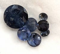 Close-up view of seven round faceted blue sapphires in various sizes on white background, showing natural inclusions and color variations.