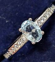 Close-up of silver ring with an oval faceted light blue topaz center stone in four prongs, featuring smaller clear cubic zirconia stones embedded along the band.