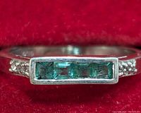 Close-up photo of the silver ring showing the three rectangular emerald stones set horizontally with accent stones on the band against a red background.