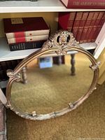 Full view of the oval vintage mirror leaning against a bookshelf showing the carved floral and crest details at the top of the frame.
