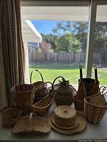 Overview of multiple assorted hand woven wicker baskets in natural light brown tones placed on a table by window with outdoor backyard background.