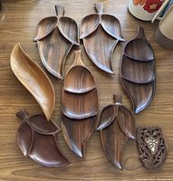 Nine wooden items including eight Monkey Pod wood bowls shaped like pods, leaves, and abstract forms laid flat on a wooden surface.