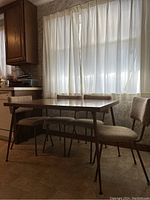 Rectangular wooden table with brown metal angled legs and four matching chairs with beige fabric upholstery in front of a window with white curtains.