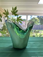 Front view of green translucent glass vase with irregular flared mouth and rounded base, set against a backdrop of green plants and window light.
