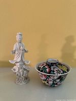 Porcelain statue standing next to floral decorated pot with lid