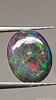 Close-up photo of oval cabochon black opal gemstone held by tweezers showing bright rainbow play-of-color with dominant green, red, blue hues.