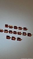 16 princess cut padparadscha sapphires arranged in two rows on white background showing color and clarity.