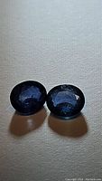 Pair of dark blue oval cut sapphires on a neutral background showing color and facets.