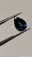 Pear-shaped deep royal blue sapphire gemstone held by tweezers. The stone is transparent with an intense royal blue color visible under the light.