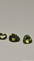 Three natural apple green peridot gemstones shown side by side on a gray background. The cuts visible are oval, pear, and trilliant. High clarity and luster visible.